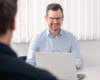 Symbolbild für den individuellen Nutzen unserer SAP-EWM-Schulungen: Ein SAP-Experte sitzt schaut lächelnd die gegenüber sitzende Person über den Laptop-Bildschirm an.